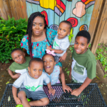 An African American woman smiles up at the camera surrounded by 5 young kids.